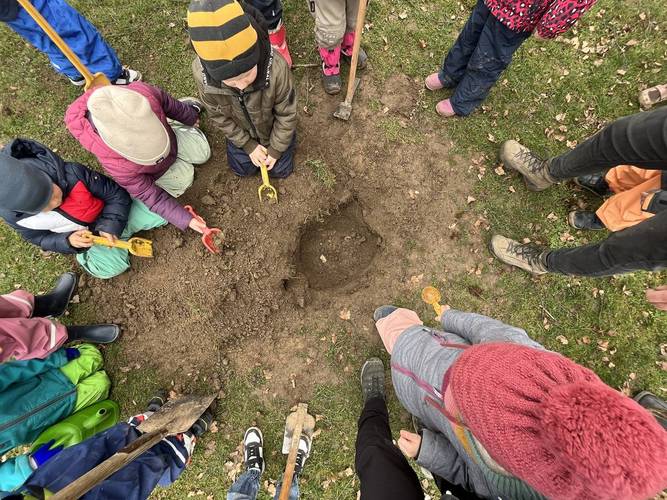 Foto der Pflanzaktion aus der Perspektive von oben. Die Kinder spielen mit kleinen Schaufeln im gegrabenen Loch