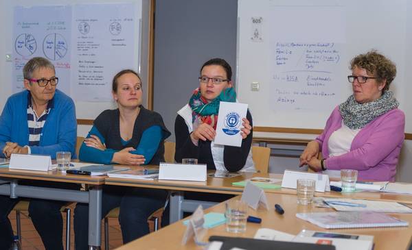 Foto eines Workshops zu nachhaltiger Beschaffung mit vier Personen an U-förmig angeordneten Tischen vor Postern an der Wand